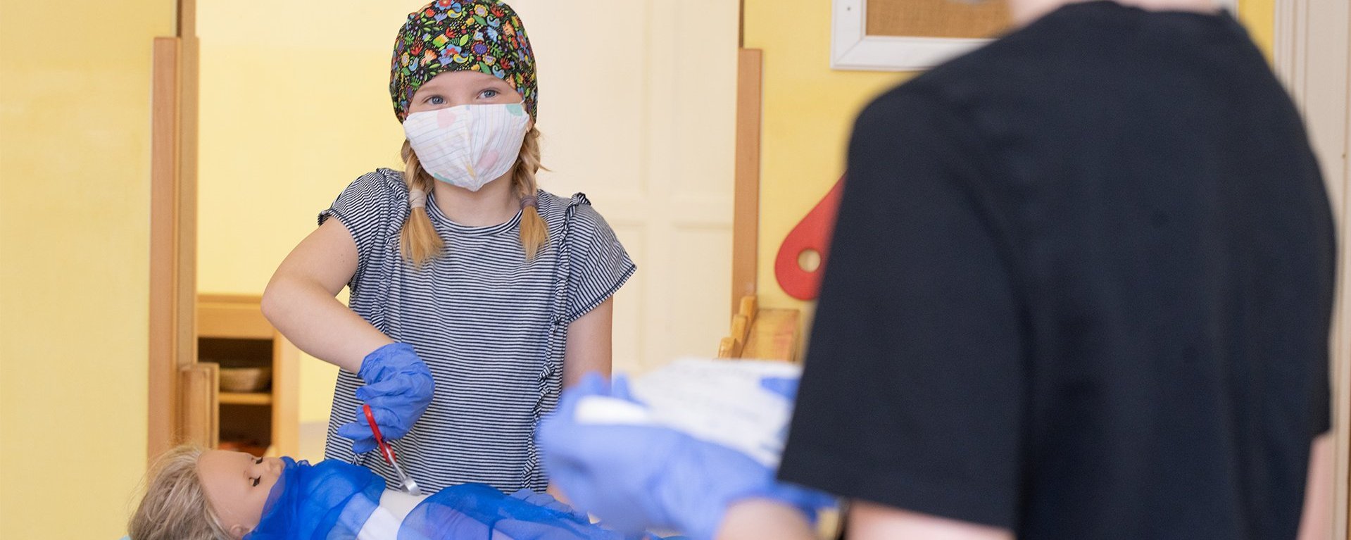 Ein Mädchen mit einer Arzthaube auf dem Kopf und einem Mundschutz operiert mit Spielzeug eine Puppe die vor ihr, mit einem Tuch abgedeckt, auf dem Tisch liegt. Das Mädchen schaut ein Kind an, welches ihr gegenüber steht.