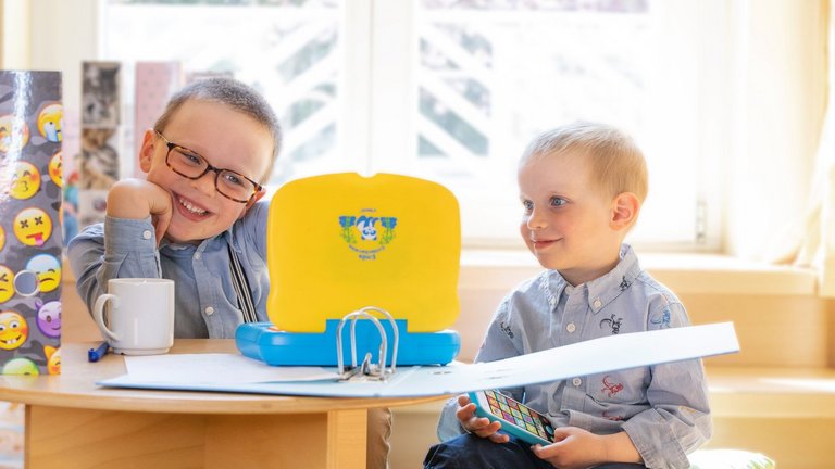Zwei Jungen sitzen hinter einem kleinen Tisch. Einer der Jungen hat braunes Haar und eine Brille auf, der andere Junge hat blondes Jahr und ein Spielzeughandy in der Hand. Auf dem Tisch stehen eine Tasse, ein Spielzeuglaptop und ein aufgeschlagener Ordner.