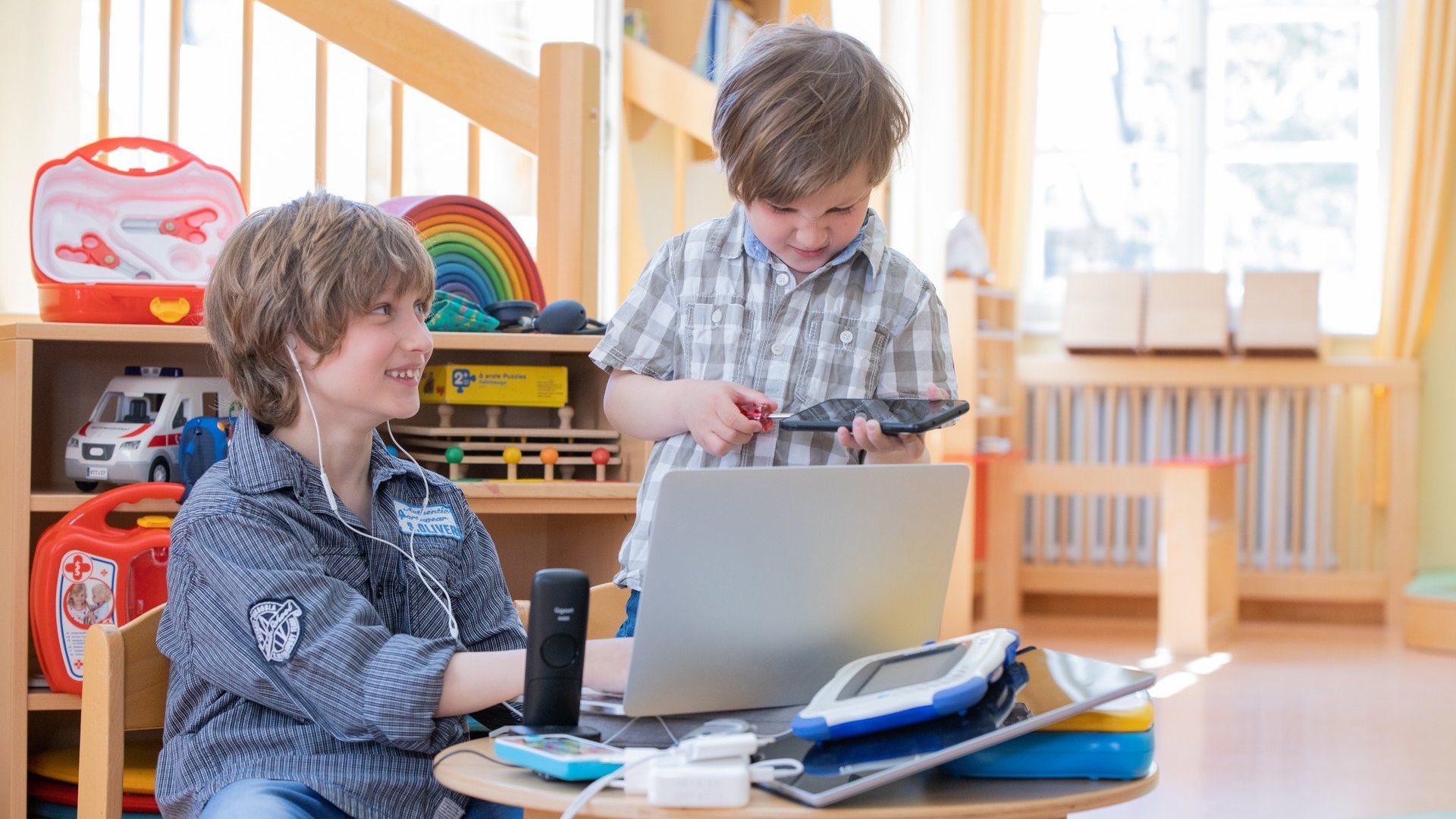 Auf dem Bild sind zwei Jungen mit braunen Haaren. Einer der beiden sitzt an einem kleinen Tisch und hat Kopfhörer in den Ohren. Er arbeitet an einem Laptop, der auf dem Tisch steht. Auf dem Tisch liegt weitere technische Gegenstände, wie z. B. ein Telefon. Der andere Junge steht neben dem Tisch und repariert mit einem Schraubenzieher ein Tablet.