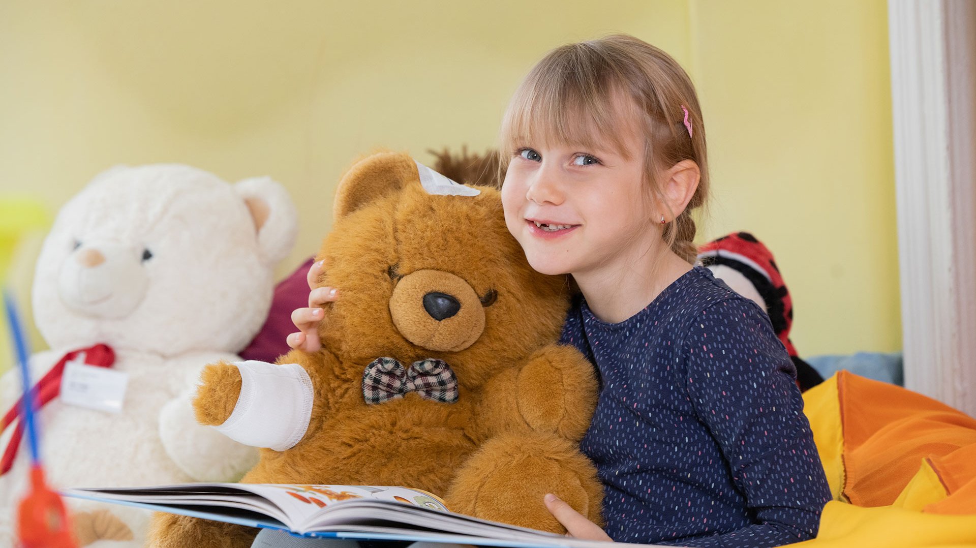Ein blondes Mädchen hat einen großen Teddybären im Arm und ein Buch auf dem Schoß. Der Teddy hat einen Verband am Arm und ein Pflaster auf dem Kopf. Das Mädchen schaut lächelnd in die Kamera.