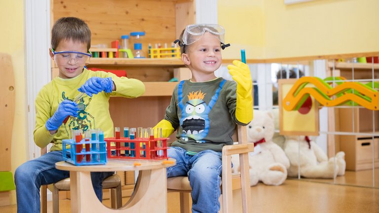 Zwei Jungen sitzen in einem Spielzimmer und haben eine Schutzbrille auf. Vor ihnen steht ein Tisch mit Spielzeug-Reagenzgläsern.